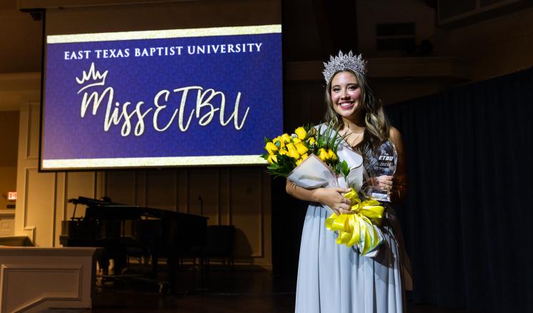 r61_1518 A female inside smiling in a ballgown with a crown on and flowers and an award in her hands with a Miss ETBU slide in the background