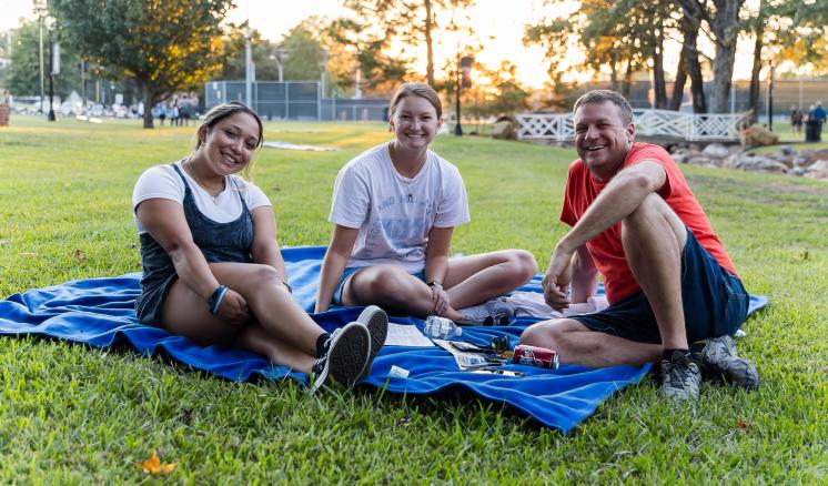 Group of two females and one male sitting on a blanket in the grass outside