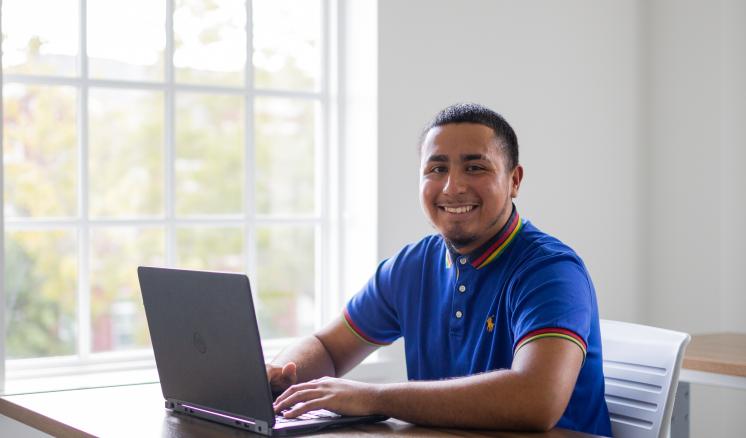 A male student sitting with laptop in a classroom by a window.