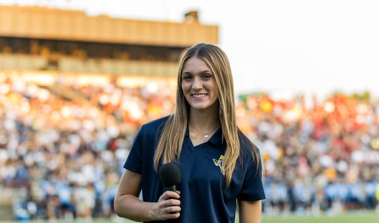 A female student holding a microphone on a football field with the stands full of fans in the background.