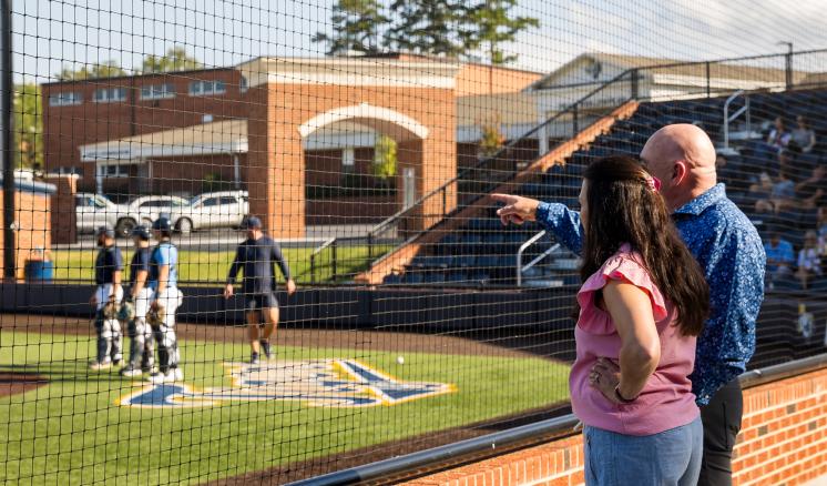 Man and woman watching and pointing to baseball team playing on baseball field.