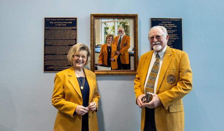 Man and woman wearing gold jackets standing in front of wall portrait smiling at the camera.