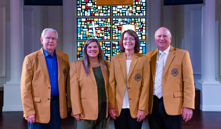  Four people in golden-beige blazers standing in front of a stained-glass window.