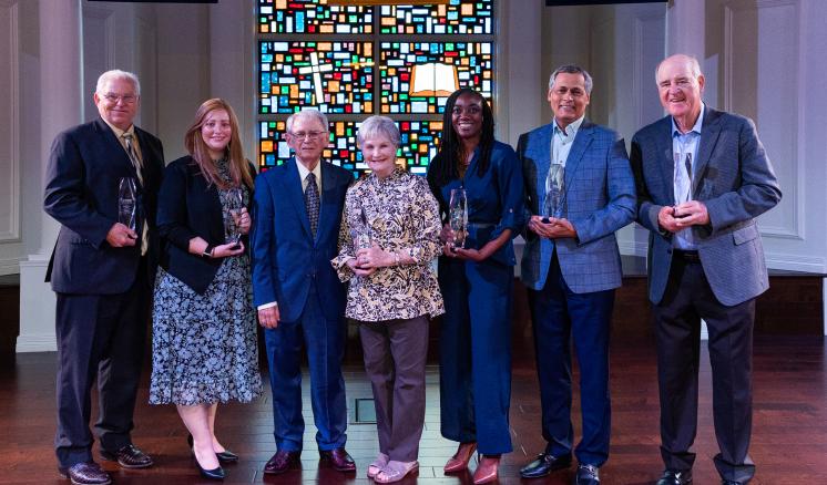 2023_homecoming_chapel_0922 A group of seven people holding awards, standing in front of a colorful stained glass window.