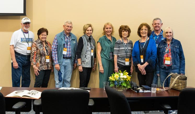 A group of nine people standing side by side in a meeting room, all wearing conference badges.