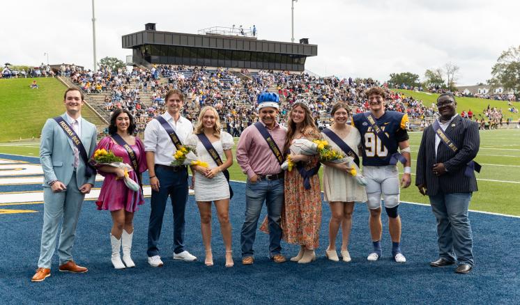  A group of nine people posing on a sports field during an event, some holding bouquets and wearing sashes.