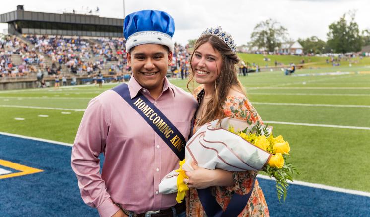 Man and woman wearing crowns and sashes stand on a football field; the man holds "Homecoming King" sash and the woman holds flowers.