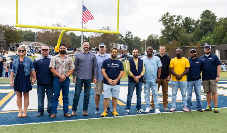 A group of eleven people standing in a line on a football field under a goalpost with an American flag in the background.