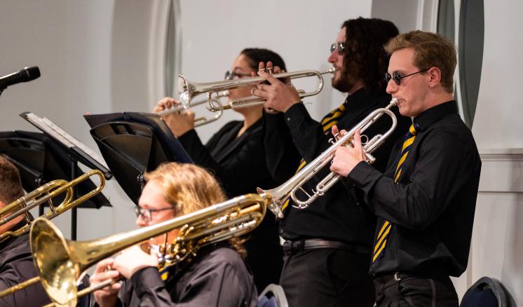 Four musicians playing trumpets and trombones in a performance, wearing black shirts and striped ties.