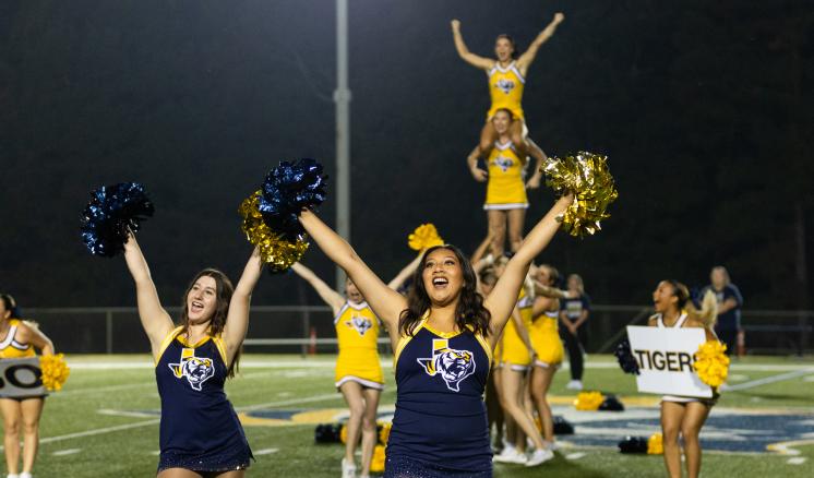 Cheerleaders with arms up on a football field in navy and gold uniforms.