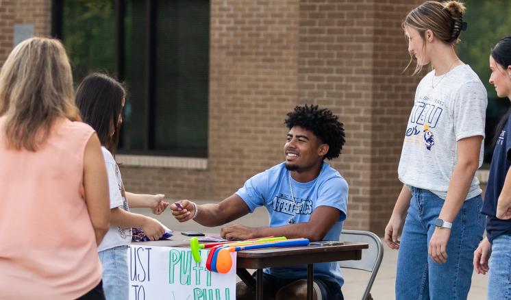 A male and female student giving a ticket to a girl at a table with a poster on the front of it