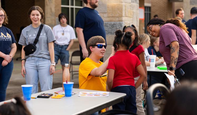 A group of male and female students at a table face painting a little girl