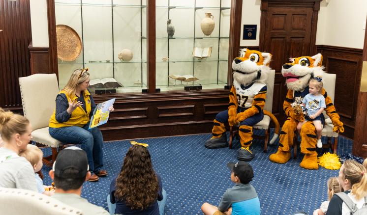 r62_9566 A woman reading a book to a group, with two tiger mascots and a child seated next to her. People seated on the floor listen attentively.
