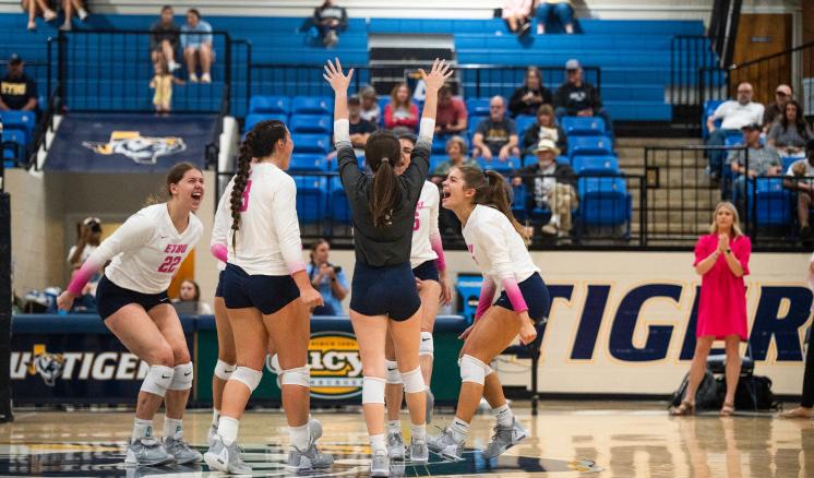 : Five female volleyball players celebrating on a gymnasium court.