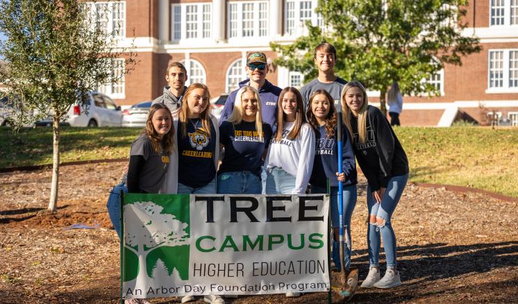 2023_arbor_day_0086 A group of nine people stand around a sign that reads "TREE CAMPUS Higher Education Arbor Day Foundation" with a brick building in the background.