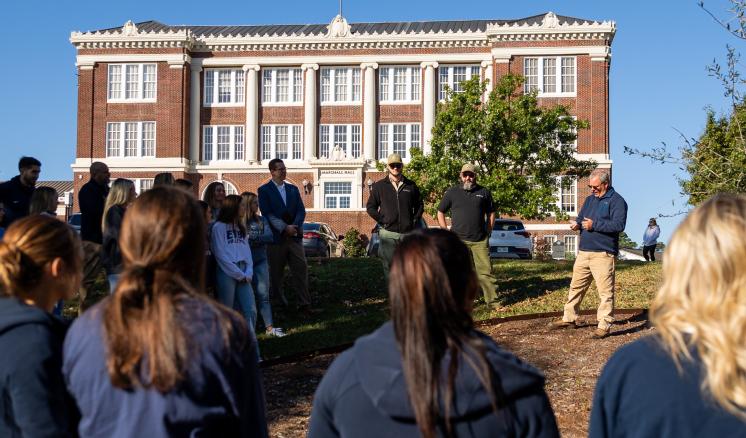 A group of people standing in front of a large brick building, listening to a speaker.