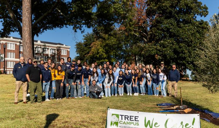  A large group of people posing outdoors with a banner that reads, "TREE CAMPUS USA - We Care! Planting for Tomorrow...Today," and trees and a brick building in the background.