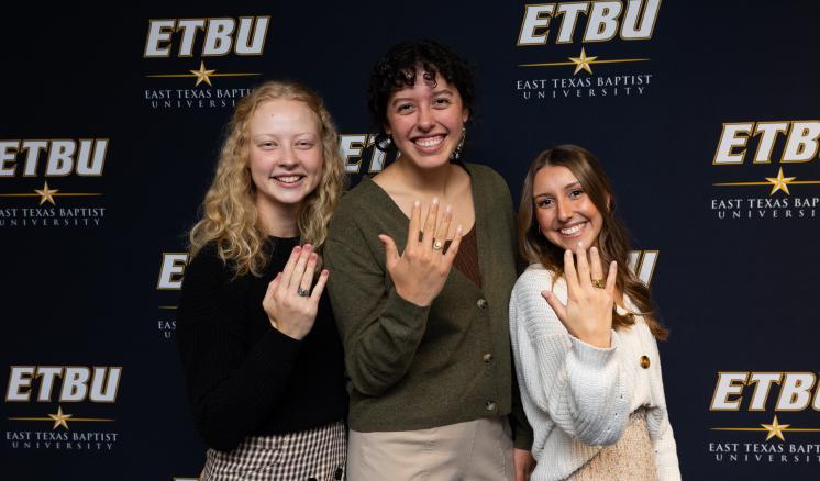 Two female students holding their hands up in front of an East Texas Baptist University navy backdrop.
