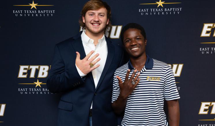 Two male students holding their hands up in front of an East Texas Baptist University navy backdrop.