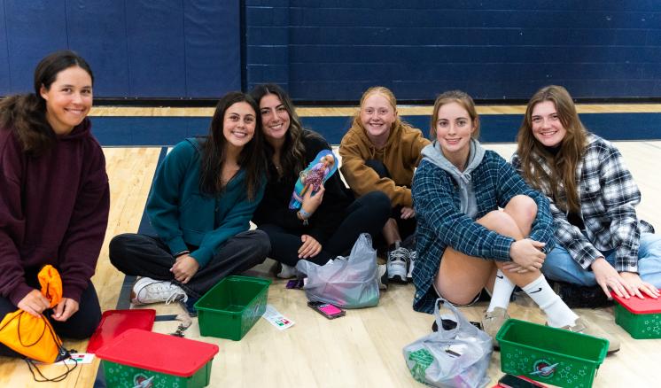 Group of female sudents sitting on the ground filling their Operation Christmas Child boxes smiling at the camera 