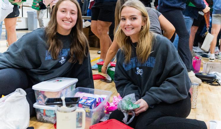 Two female students sitting on the ground in gymnasium filling boxes for Operation Christmas Child 