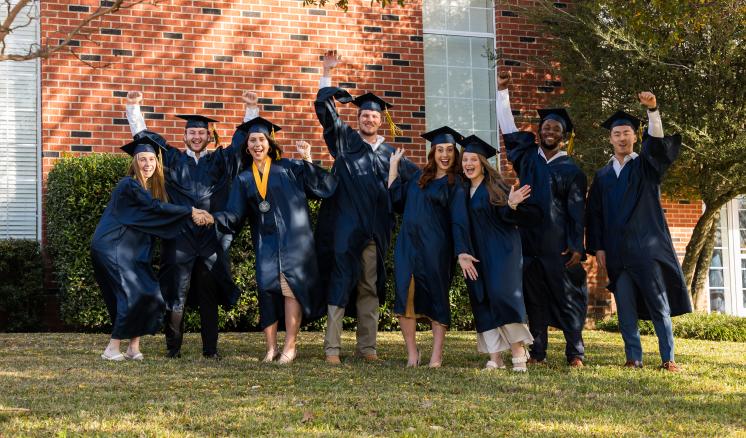Group of male and female students in navy cap and gown smiling at the camera with arms in the air 