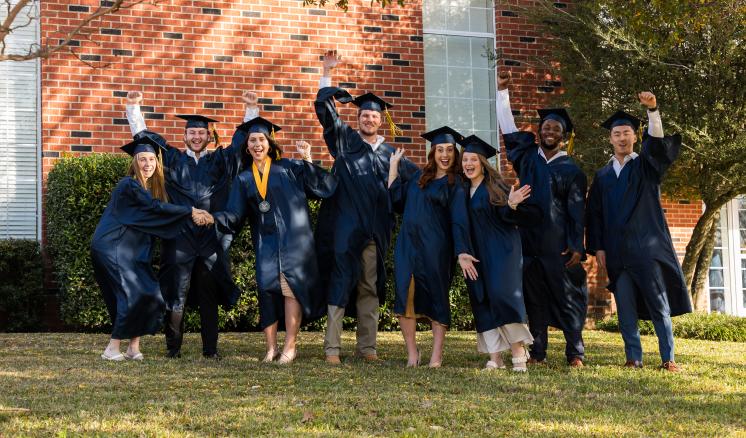Fall 2023 Commencement Group of male and female students in navy cap and gown smiling at the camera with arms in the air