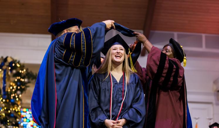 Female graduate student in navy cap and gown being hooded by faculty members in academic regalia