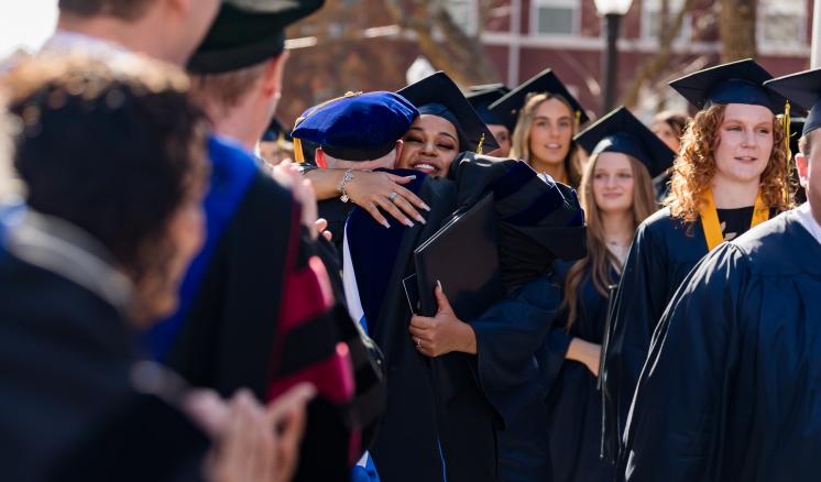 Female student in cap and gown hugging professor outdoors 