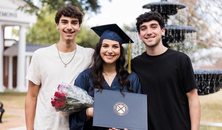 A female graduate holding a diploma smiling with two men in front of water fountain.