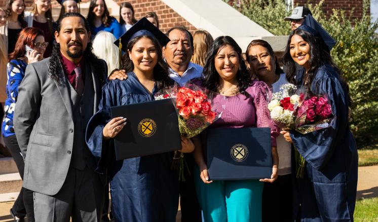 Two graduates holding their diplomas posed with family outdoors smiling at the camera 