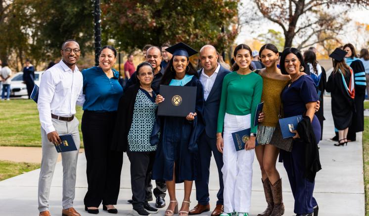 Fall 2023 Commencement Graduate in navy cap and gown posed with family outdoors smiling at the camera