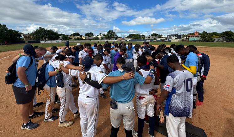 Baseball players praying in a circle with their arms around each other outdoors on a baseball field