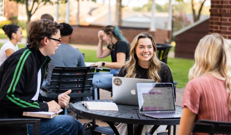 Group of three students talking to each other while sitting at a table outdoors 