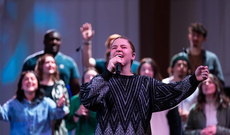 Female student singing into a microphone on a stage with a choir behind her 