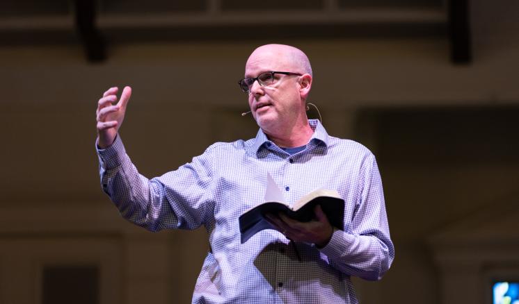 2024_spiritual_renewal_monday_chapel_0478 Man speaking on a stage with a Bible in his hand