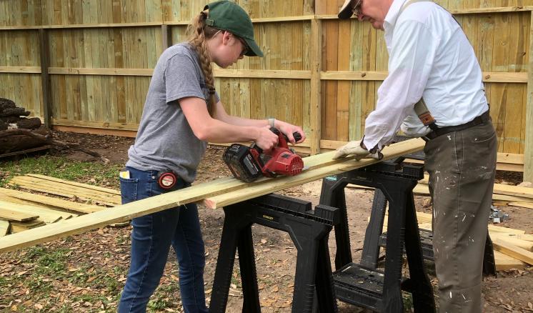 Woman operates a table saw to cut a piece of wood held up between two saw horses