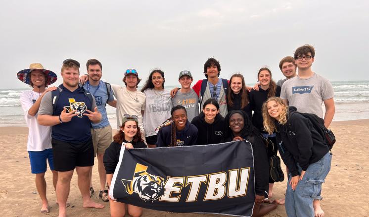 img_6567 A group of individuals pose on the beach holding a flag with the ETBU logo on it