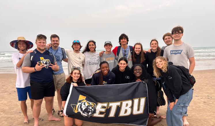 A group of individuals pose on the beach holding a flag with the ETBU logo on it