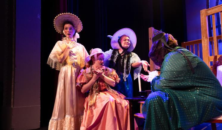 A group of actors in colorful period costumes perform a scene from a play on stage, with vibrant lighting highlighting their expressions.