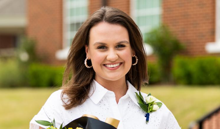 Girl in a white dress standing in front of a red brick building holding a bouquet of yellow roses.