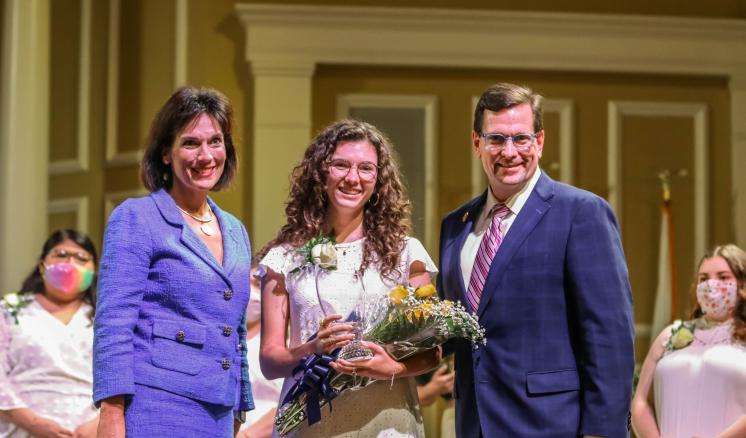 An older woman, a college female holding an award and yellow flowers, and an older man smiling at the camera.