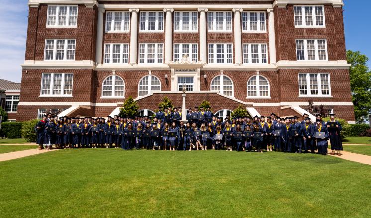 A group of college graduates in navy caps and gowns hold their degrees in front of a red brick building