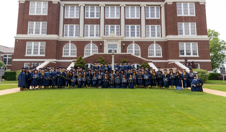 A group of college graduates in navy caps and gowns hold their degrees in front of a red brick building