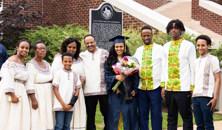 A large family surrounding a graduate in a navy cap and gown holding flowers in front of a red brick building.