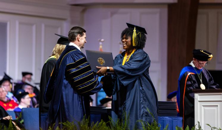 During a commencement ceremony, a woman in a navy cap and gown on the right receive her diploma from a man in navy academic regalia on the left.