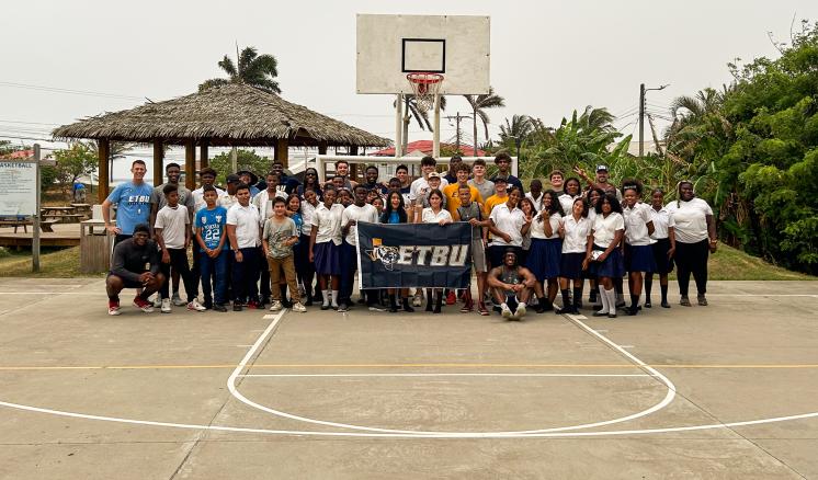 ETBU Men's Basketball Team stand on a basketball court with youth in Honduras holding an ETBU-branded flag