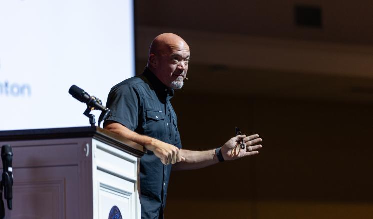 A male speaker standing behind a podium, gesturing with his hands while speaking to an audience. 