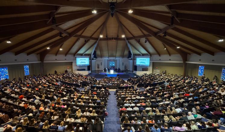 A large congregation inside an auditorium, filled with people seated in rows, facing a stage with a speaker.