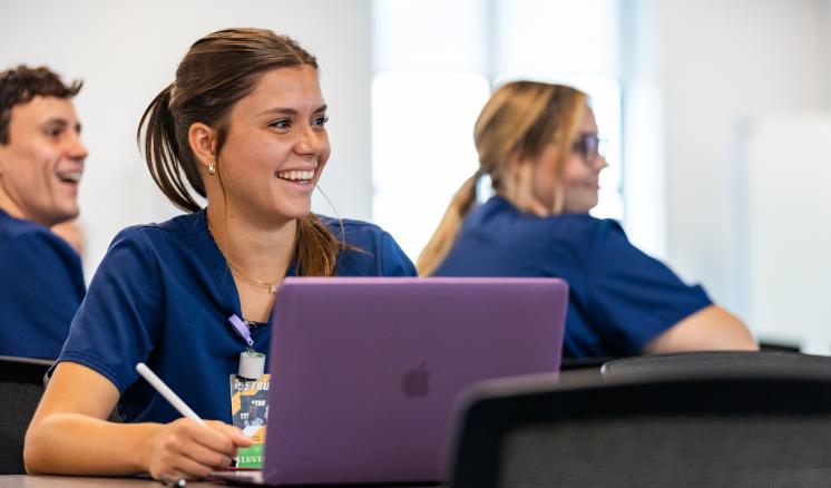 A young woman, dressed in blue scrubs, is seated in a classroom, smiling while working on a laptop. She holds a stylus in one hand, with other students visible in the background. 